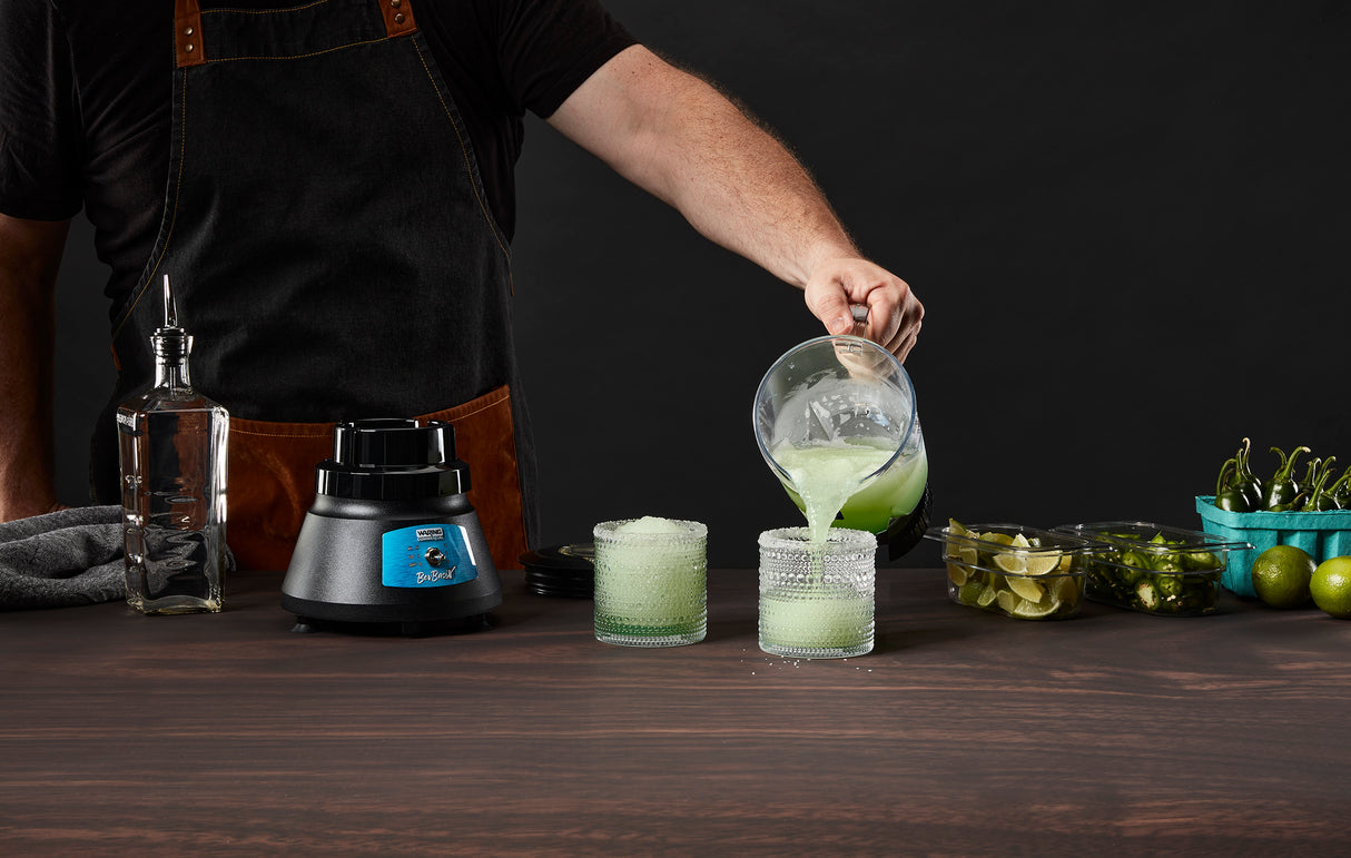 A person in a black shirt and apron pours a green blended drink from a pitcher into two textured glasses on a wooden table, surrounded by a Bar Blender with stainless steel blade, limes, sliced peppers, and containers of garnish. Background is dark.