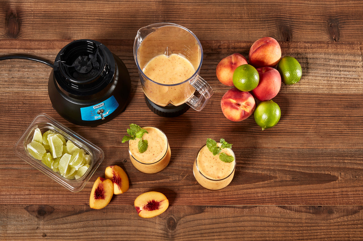 A bar blender with a copolyester container sits on a wooden table beside limes, lime wedges, whole and sliced peaches, and two glasses of smoothie garnished with mint. The ingredients are arranged neatly around the blender.