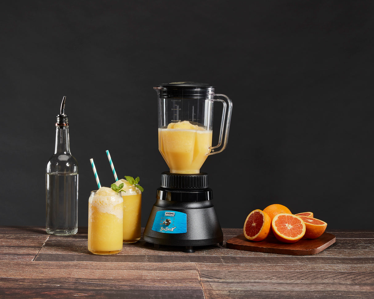 A 48 oz. capacity Bar Blender filled with orange slush sits on a wooden table. Nearby are a glass bottle with a spout, two garnished glasses of slush, and a cutting board with whole and halved blood oranges against a dark background.