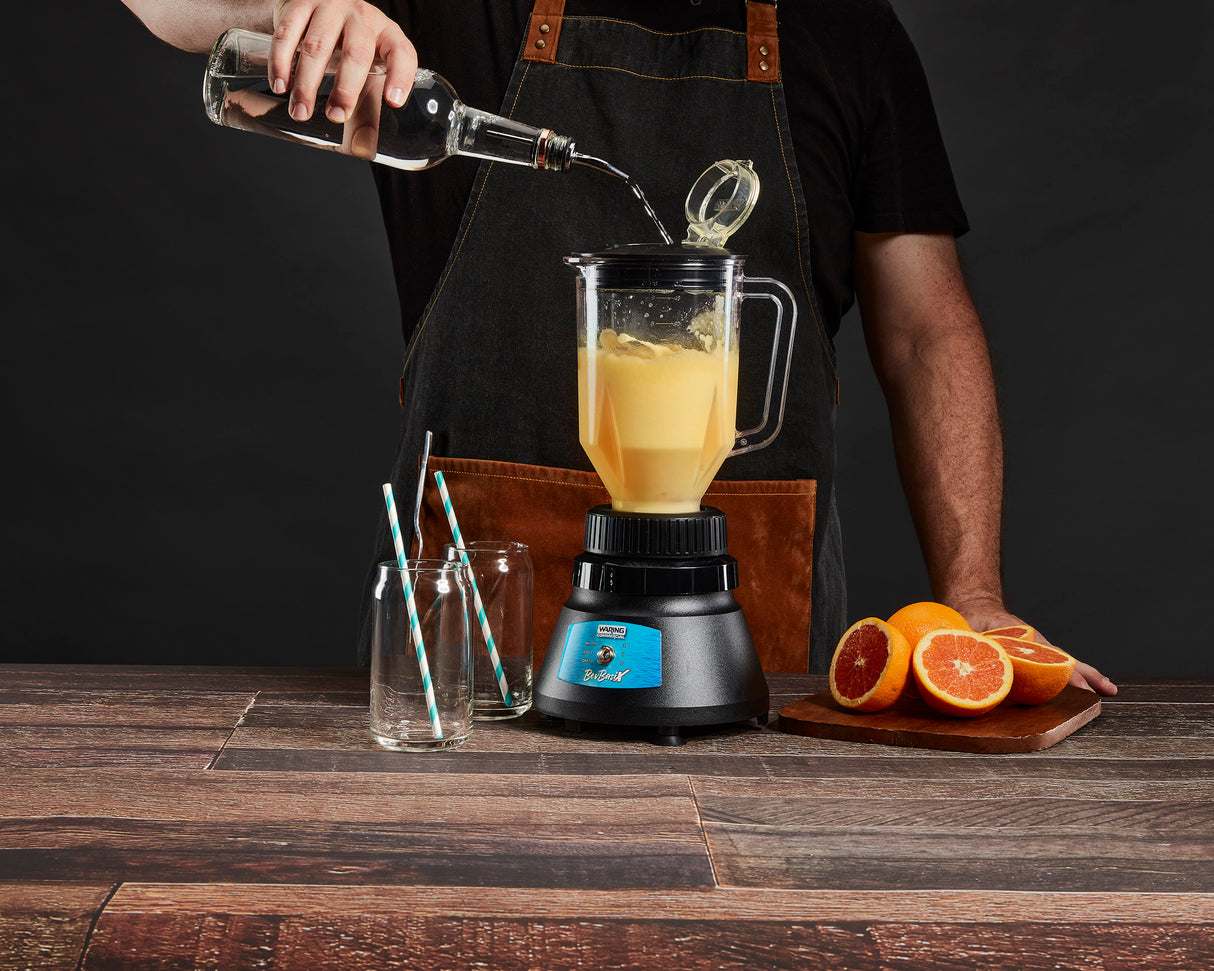 A person in a brown apron pours clear liquid from a bottle into a bar blender with a copolyester container filled with orange juice. Two empty glasses with striped straws and halved oranges sit on a wooden table against a dark background.
