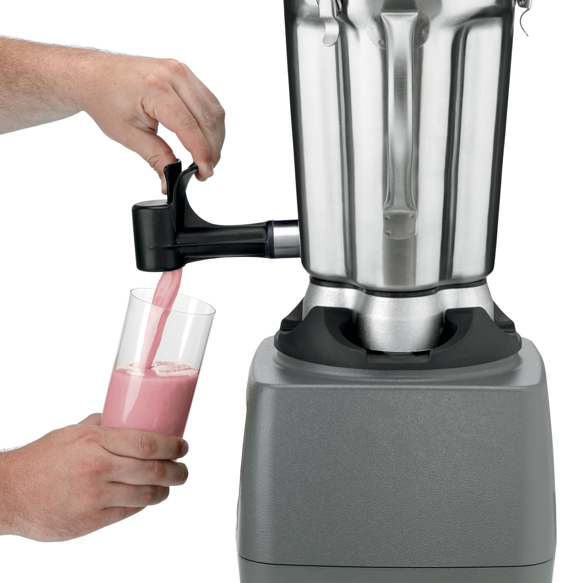 A person uses a lever spout to pour a pink smoothie from a heavy-duty blender with a stainless steel container into a clear glass. One hand operates the lever while the other holds the glass; the blender sits on a gray base against a plain white background.