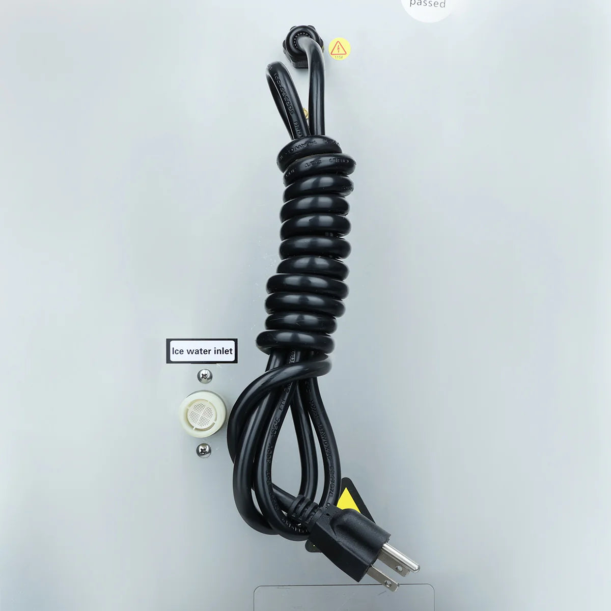 A black electrical cord neatly coiled and tied hangs from a hook on a light gray commercial ice maker. Below the cord is a labeled “Ice water inlet” port with a white cap, and above, a small yellow warning triangle symbol is visible.