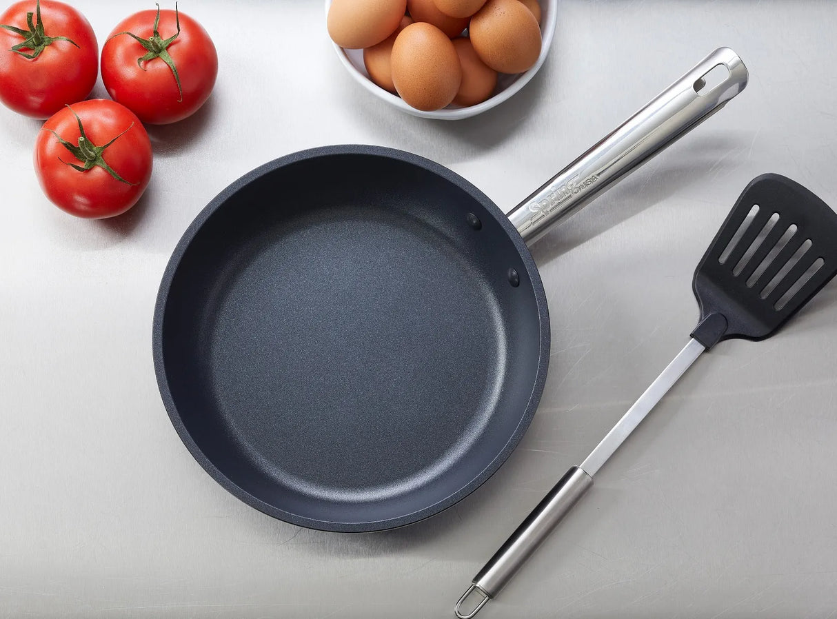 An Endurance Fry Pan sits on a stainless steel surface next to a black spatula, a bowl of brown eggs, and three red tomatoes with green stems—neatly arranged for an induction cooking preparation scene.