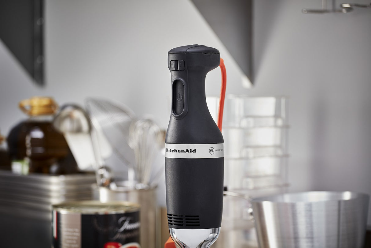 A close-up of a black KitchenAid immersion blender stands upright in a modern kitchen, surrounded by various utensils, a whisk, stacked containers, a tin can, and a bottle in the blurred background.