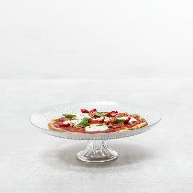 A Fortessa Jupiter clear cake stand holds a small pizza topped with melted cheese, tomato slices, pepperoni, and fresh basil, set against a light gray textured background. The scene is minimalistic with no other objects present.
