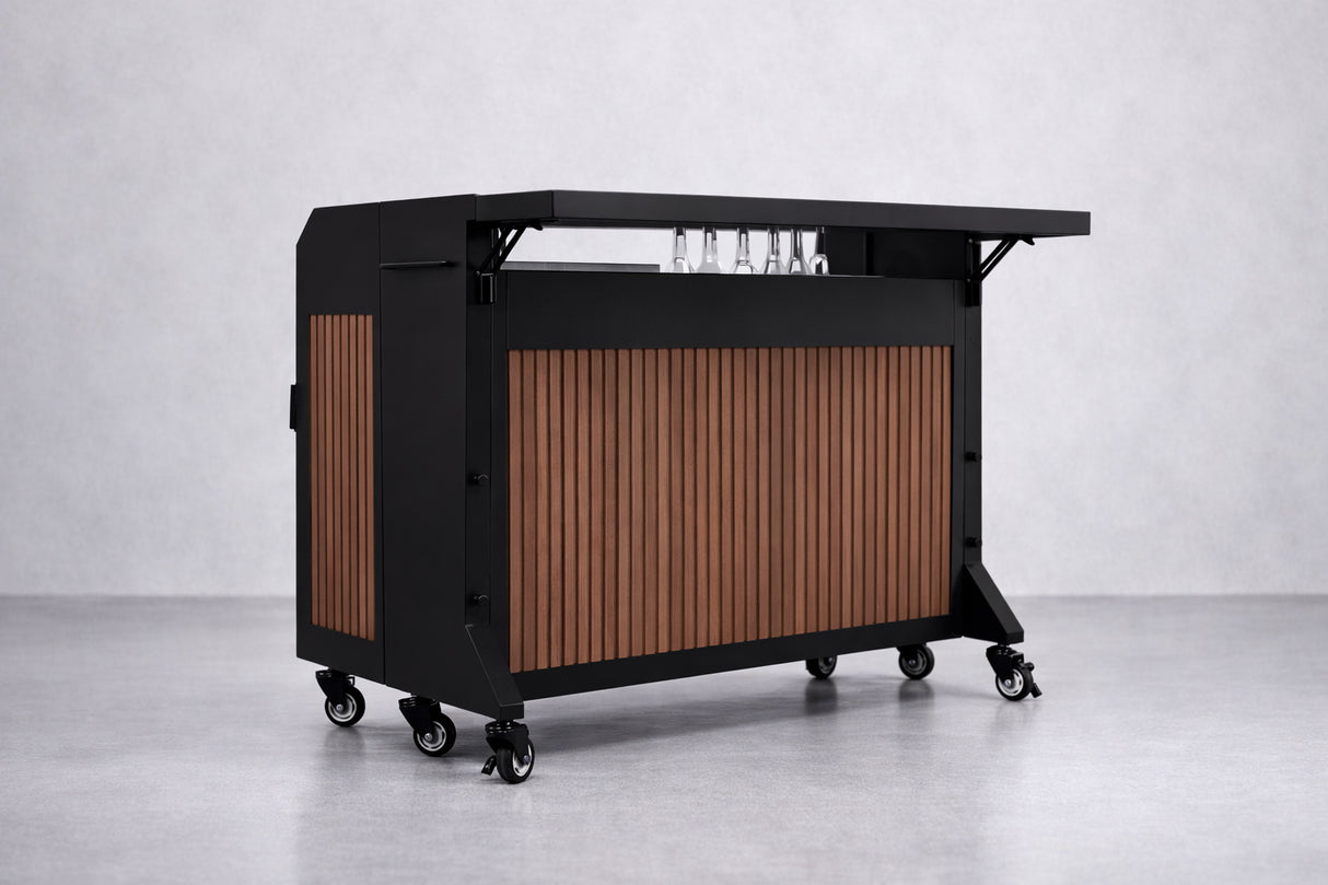 The NomadBar Mobile features a black metal frame, wood slat paneling, and a flat black countertop. With four caster wheels, glass holders, and an insulated ice bin, this collapsible bar system sits against a light gray textured wall and floor.