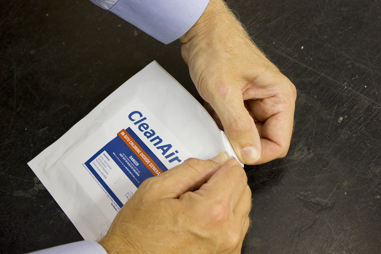 A pair of hands opens a white CleanAir Pack, an ice machine sanitizer, on a dark surface. The person peels back the sealed edge, revealing part of a blue and orange label with product details on the front.