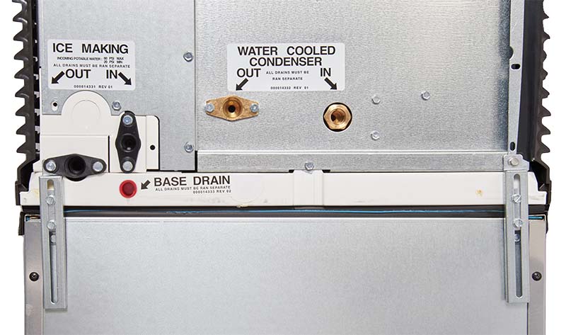 Close-up of a metal appliance panel with labeled ports: “ICE MAKING” and “WATER COOLED CONDENSER,” each with “IN” and “OUT” connections. Below, a white strip labeled “BASE DRAIN” is visible, along with adjustable stainless steel feet nearby.