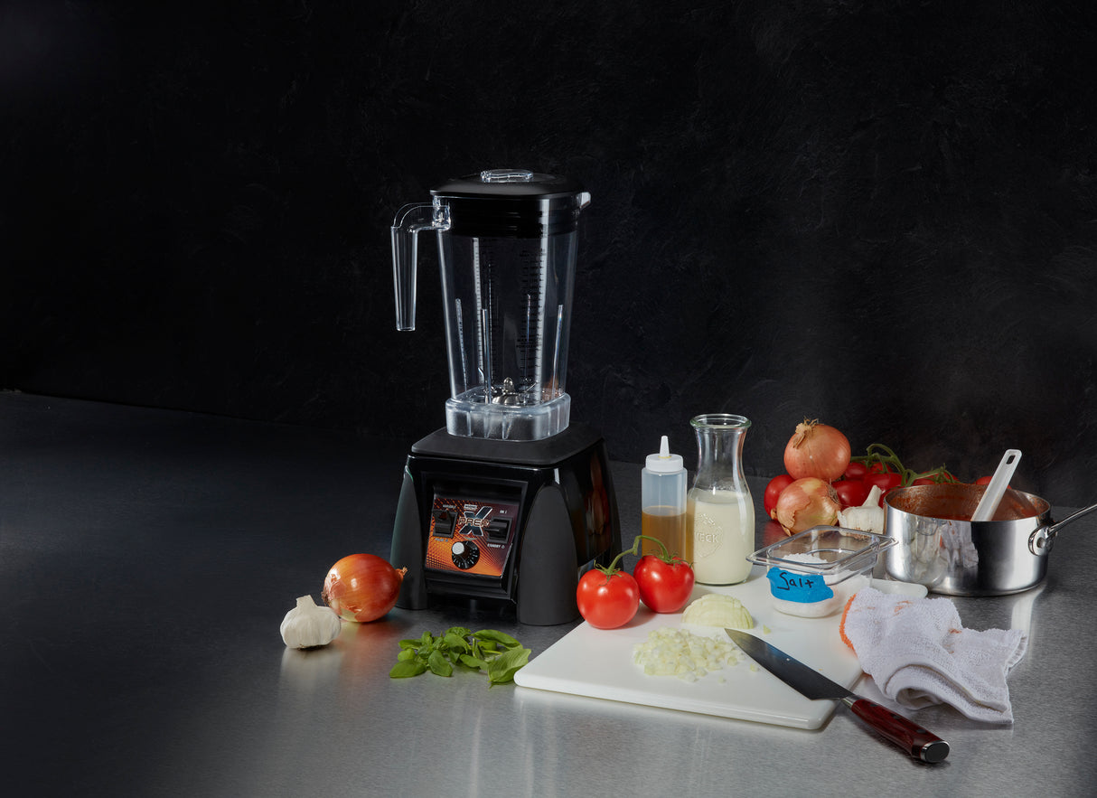 A heavy duty blender sits on a counter beside a cutting board with chopped onions and a knife. Nearby are tomatoes, fresh basil, garlic, a bottle of oil, jar of cream, onions, tomato sauce in a pot, butter, and a white towel.