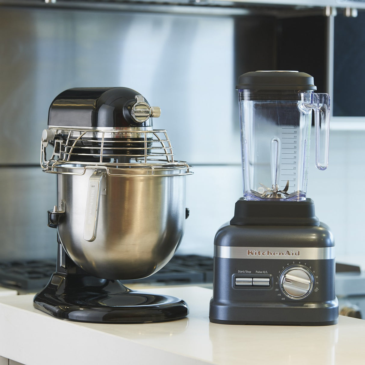 A black KitchenAid Commercial Stand Mixer with an 8 quart bowl and wire guard sits beside a gray Blender with a clear pitcher on a white kitchen countertop. A stovetop and stainless steel backsplash are in the background.