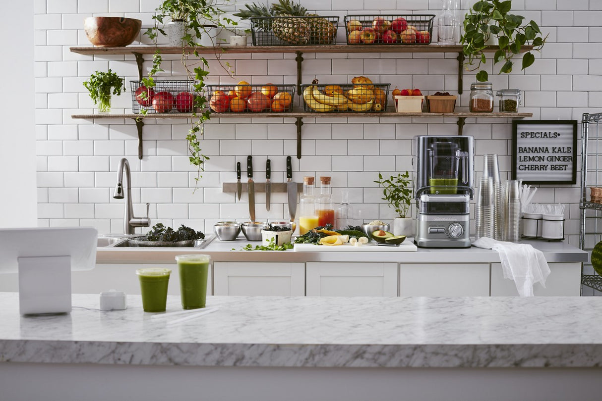 A bright kitchen with a marble counter holds two green smoothies made with a BPA free blender. Behind it are white cabinets, a juicer, knives, and assorted fruits and vegetables on wooden shelves. Potted plants and a menu board decorate the white-tiled backsplash.
