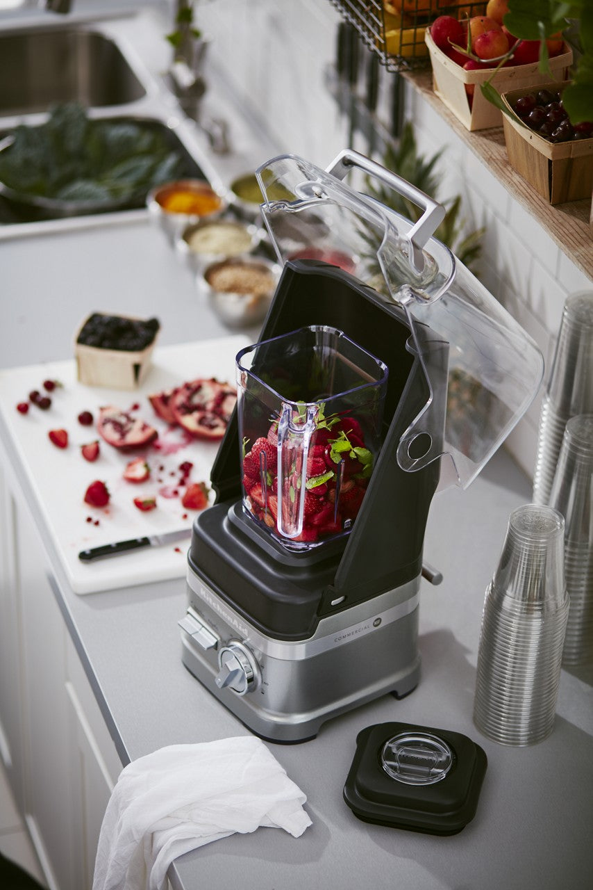 A BPA free blender with a clear sound shield is filled with chopped red fruit. It sits on a kitchen counter beside a cutting board with cut pomegranate, scattered berries, stacked clear cups, a dish towel, and baskets of fresh fruit.