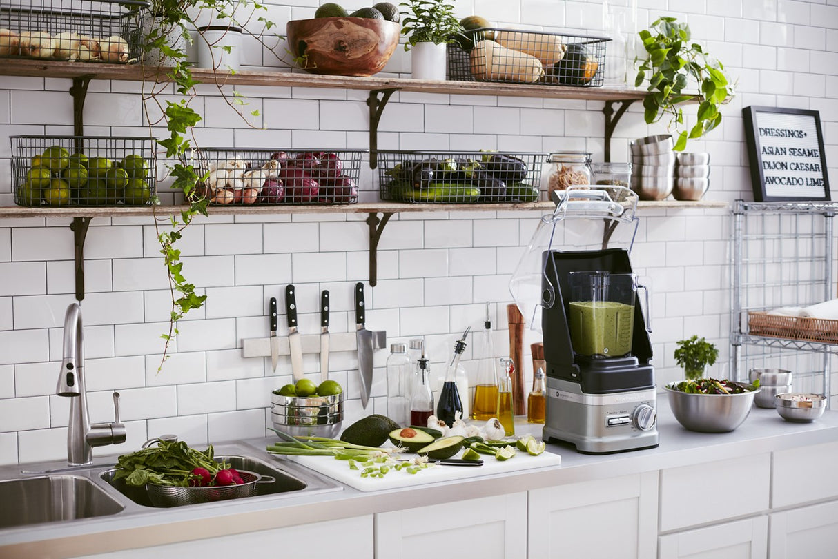 A modern kitchen with white tiled walls and a white countertop. A KitchenAid Commercial Blender filled with green smoothie sits beside fresh vegetables, knives, and bottles. Open shelves above hold produce baskets, bowls, and kitchenware. A sink is visible on the left.