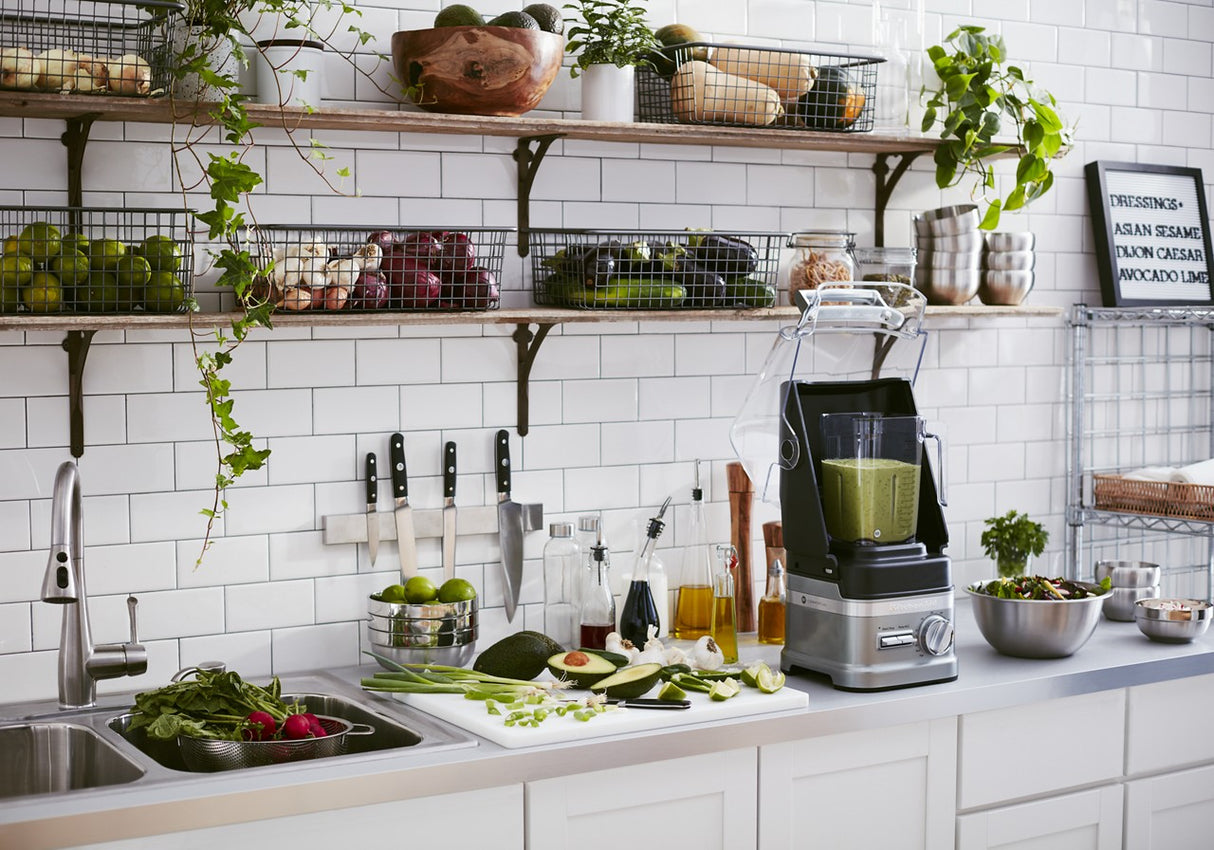 A modern kitchen with white subway tiles, open shelves holding fresh produce, plants, and kitchenware. On the countertop, a KitchenAid Commercial Blender with 60 oz. capacity blends a green smoothie beside vegetables, salad, and glass bottles near the sink.