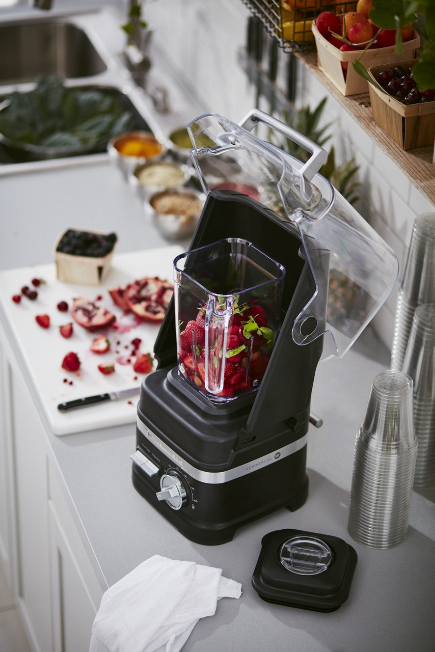 A KitchenAid Commercial Blender with a clear, open lid contains chopped red and green fruits on a kitchen counter. Nearby are stacked plastic cups, a cutting board with pomegranate and berries, and baskets of fresh fruit on a shelf.