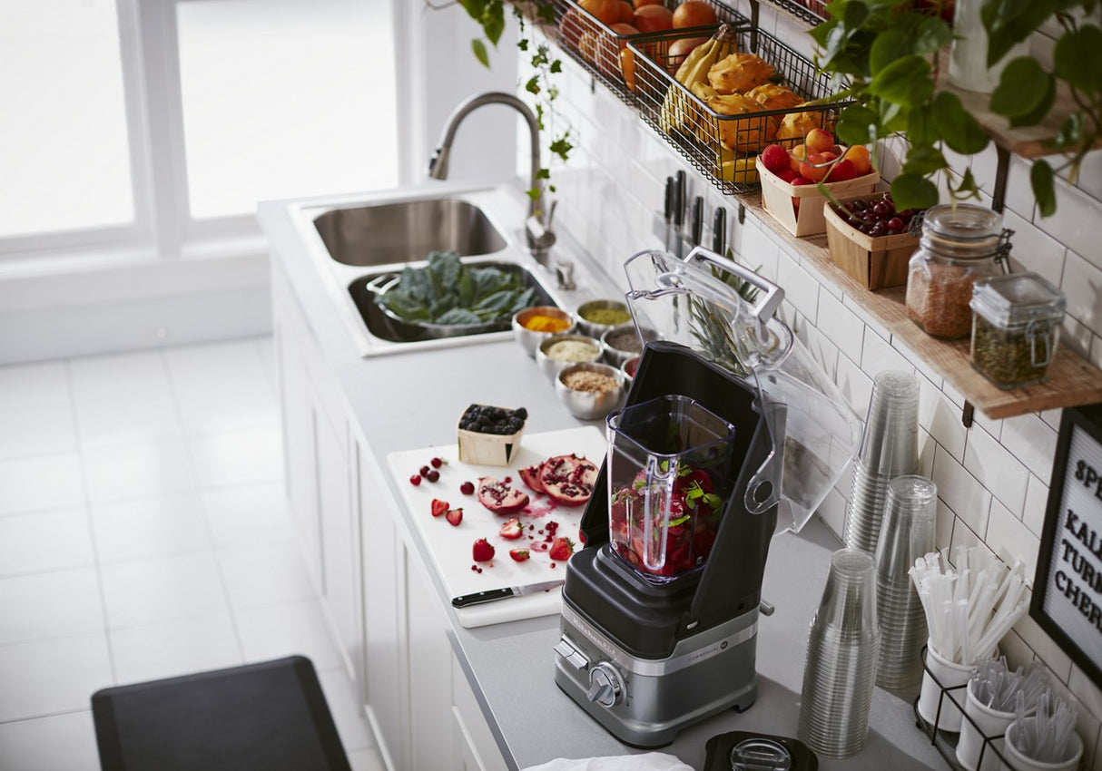 A modern kitchen countertop features a KitchenAid Commercial Blender with a 60 oz. capacity, mixing berries and pomegranate seeds. Nearby are sliced fruit, greens, jars, bowls of ingredients, stacked cups, and open shelves against a white tile backsplash.