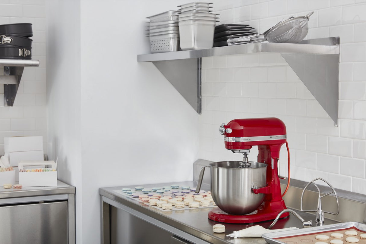 A modern kitchen with white tile walls and stainless steel counters. A KitchenAid Commercial Stand Mixer sits on the counter near rows of colorful macarons. Above are metal shelves with stacked pans, trays, and a colander.