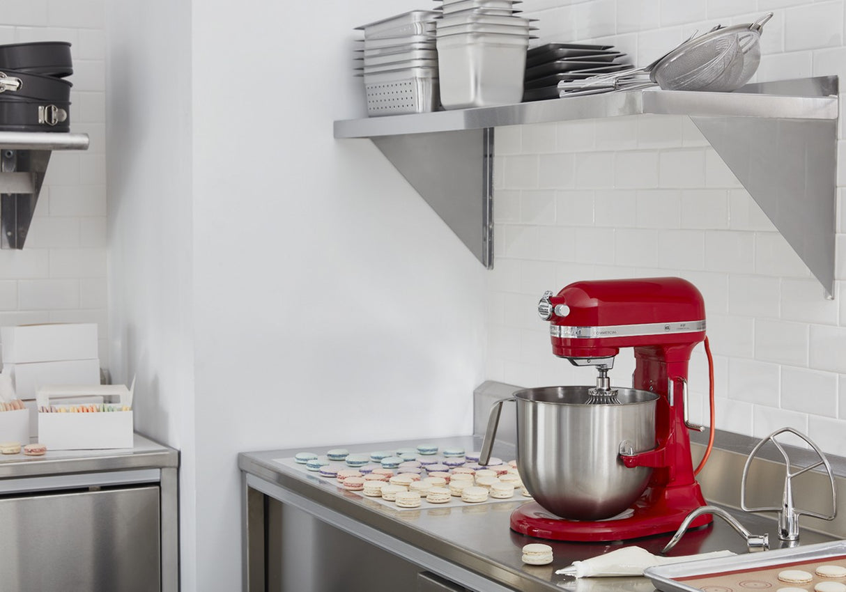 A shiny red KitchenAid Commercial Stand Mixer sits on a stainless steel countertop in a modern kitchen with white subway tile walls. Nearby, pastel macarons are arranged on parchment, while baking trays and bowls rest on open shelves above.