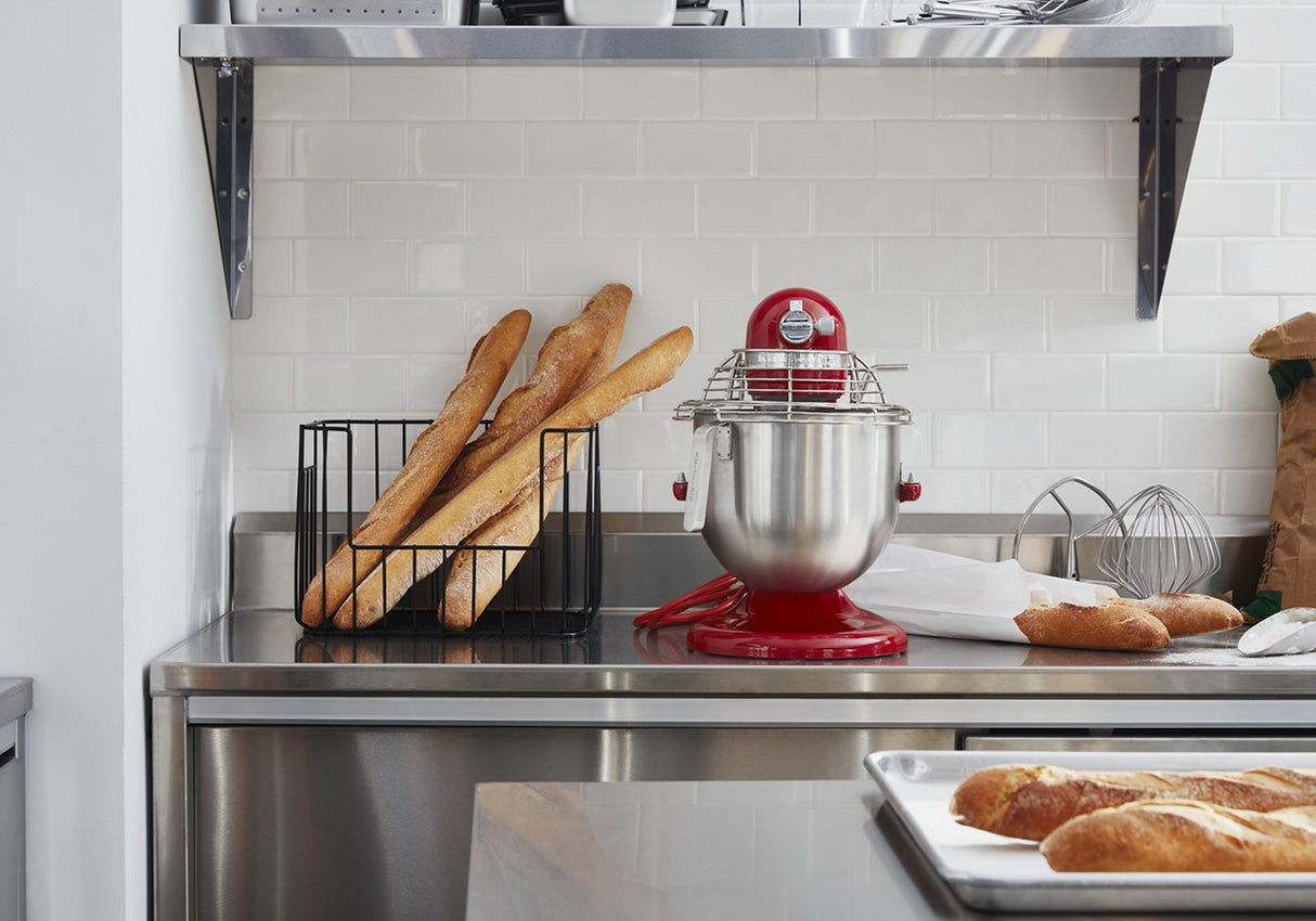 A modern kitchen counter with a KitchenAid Commercial Stand Mixer and 8 quart bowl, a metal tray of baguettes, a wire basket of baguettes, a whisk, and paper bag. The backdrop features white subway tile and a metal shelf with bowls and kitchen items.