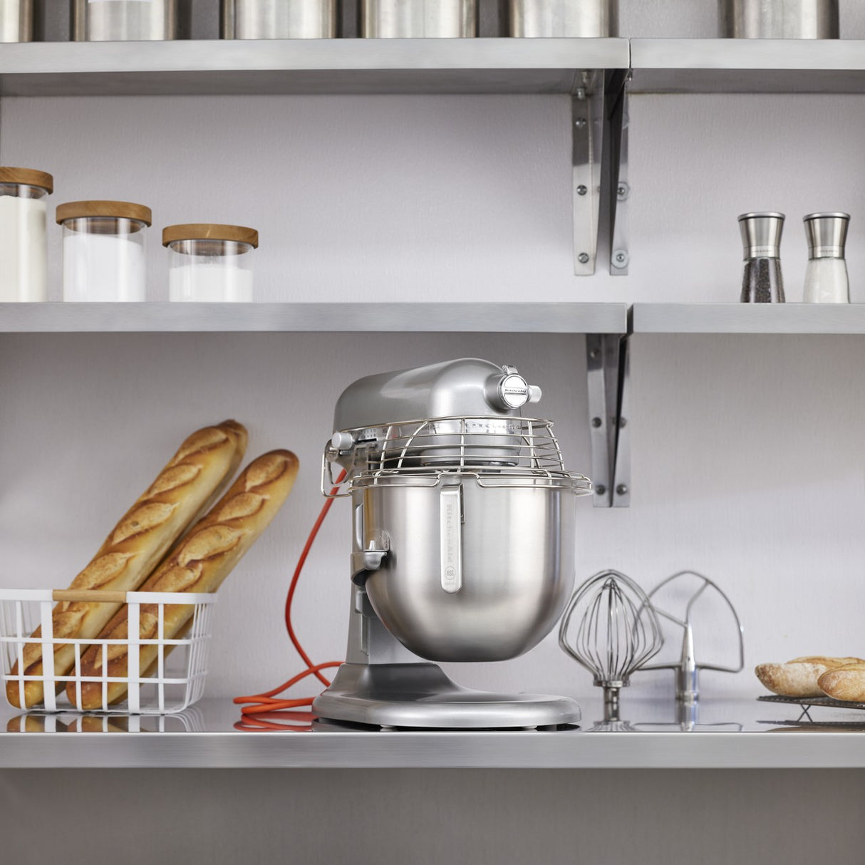 A KitchenAid Commercial Stand Mixer with an 8 quart bowl sits on a kitchen counter beside mixing attachments and a wire basket holding two baguettes. Above, shelves hold jars, canisters, and salt and pepper shakers in this clean, minimalist kitchen.