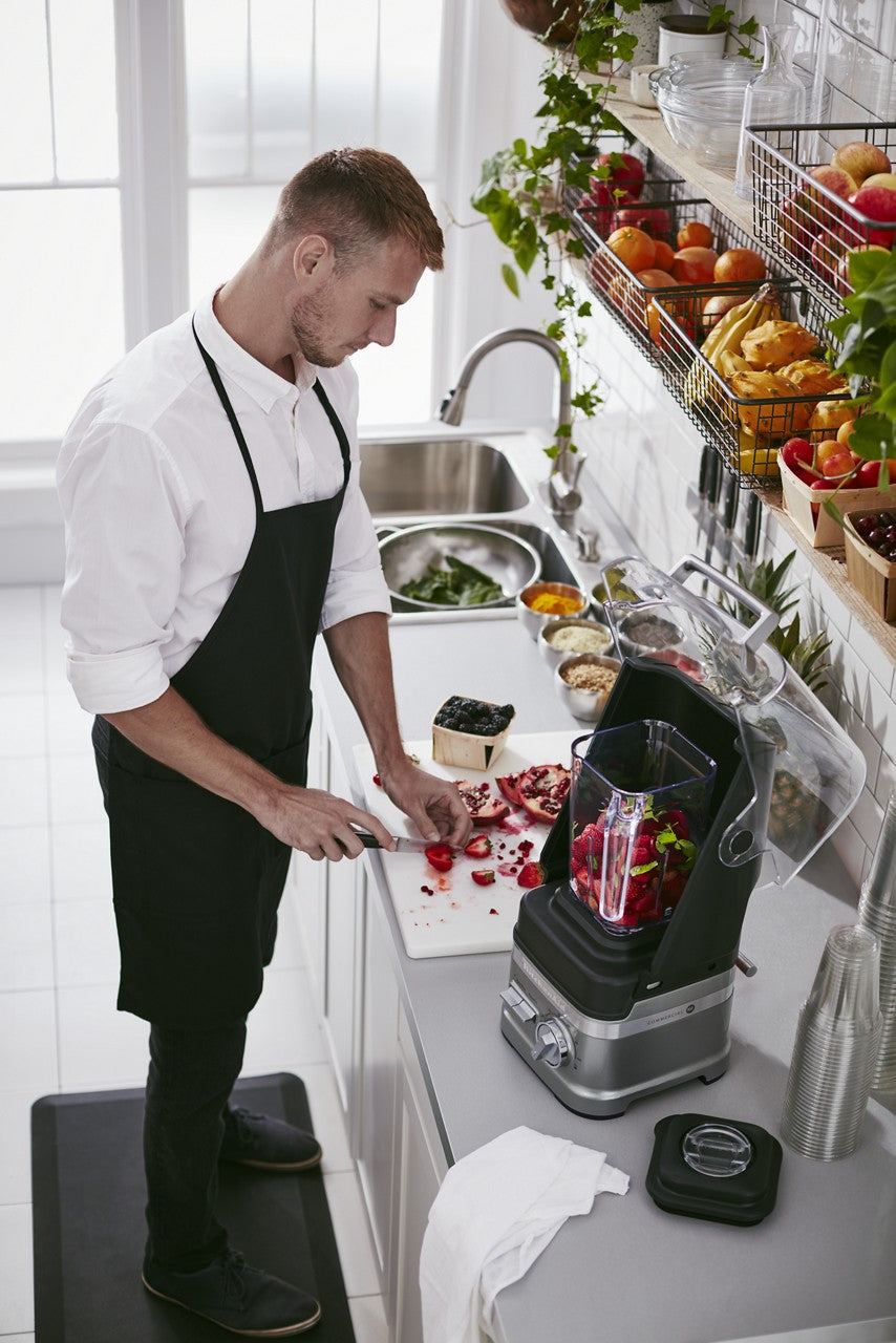 A man in a white shirt and black apron slices strawberries on a cutting board in a bright kitchen. Fresh fruit fills bowls, and a BPA free blender with berries stands on the counter. The organized kitchen is filled with natural light from large windows.