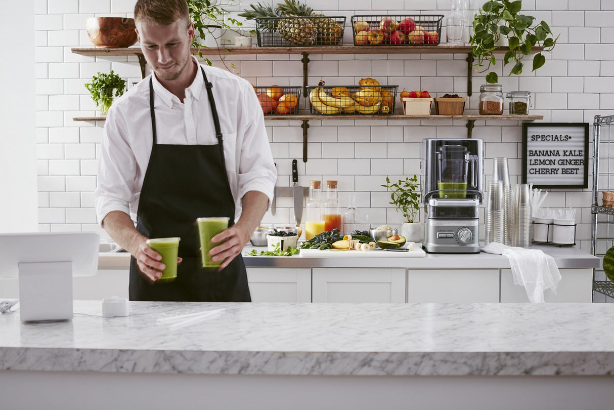 A man in a black apron stands behind a marble counter, holding two green smoothies. The kitchen features white tiles, fresh produce, and a KitchenAid Commercial Blender on the counter. A menu lists Banana Kale, Lemon Ginger, and Cherry Beet smoothies.