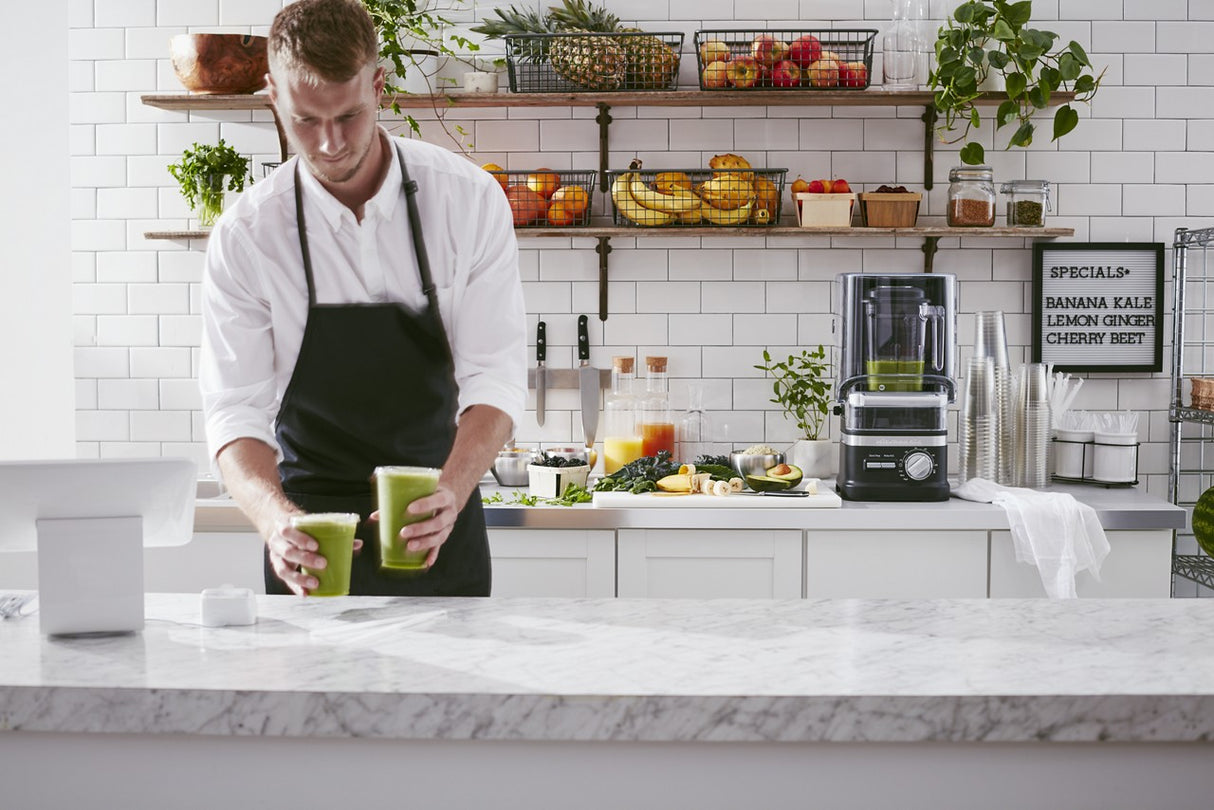 A man in a white shirt and black apron places two green smoothies on a marble counter in a bright juice bar, with fresh fruit, juices, and an Enclosure Blender with 60 oz. capacity on shelves behind him. A chalkboard menu hangs on a tiled wall.