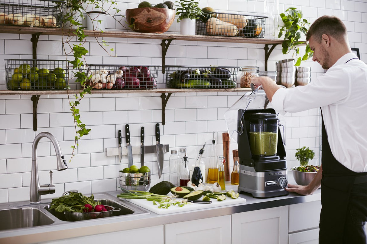 A man in an apron stands in a modern kitchen, blending green smoothie ingredients with a KitchenAid Commercial Blender. The countertop holds fresh vegetables and utensils, while open shelves display produce and jars above a white tile backsplash.