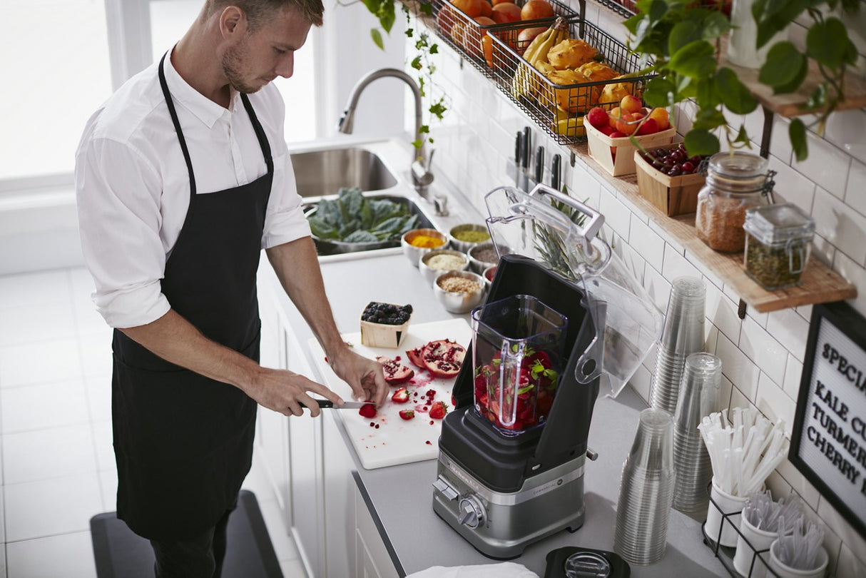 A man in a black apron slices fruit on a cutting board in a modern kitchen. He prepares ingredients for his BPA free blender, which contains chopped fruit. The counter holds bowls of fresh produce, with plants and kitchenware visible above.