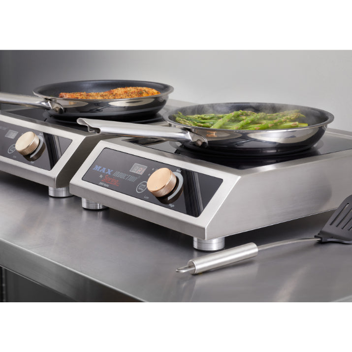 Two stainless steel induction cooktops sit side by side on a counter. One pan contains cooked salmon fillets, and the other has green asparagus. A metal spatula and digital probe thermometer rest nearby on the countertop range. Steam rises from the asparagus pan.
