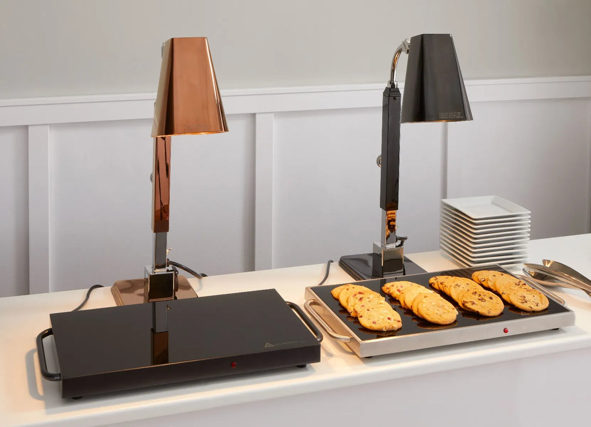 Two buffet heat lamps with a sleek stainless steel body stand on a counter. The lamp on the right shines over cookies, while the left stands over an empty tray atop a tempered glass surface. White plates and napkins complete this clean, modern setup.