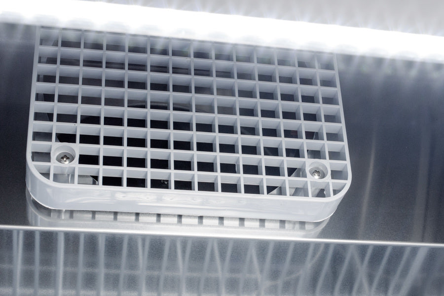 Close-up of a vent or air grille with a white plastic grid mounted on the door of a commercial refrigerator. Two screws secure it at the corners. Bright fluorescent light shines above, casting reflections and shadows on the shiny metallic surface below the vent.