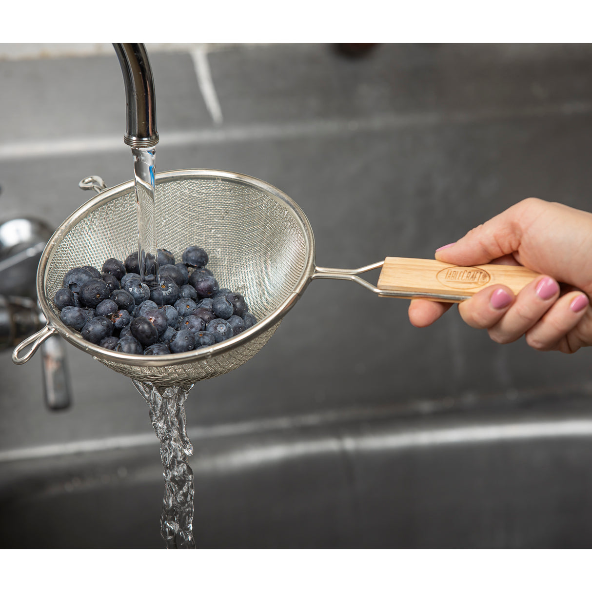 A hand holds a double fine mesh strainer with a wooden handle under running water in a sink, rinsing fresh blueberries. The water flows through the mesh, cleaning the berries. The person's fingernails are painted light pink.