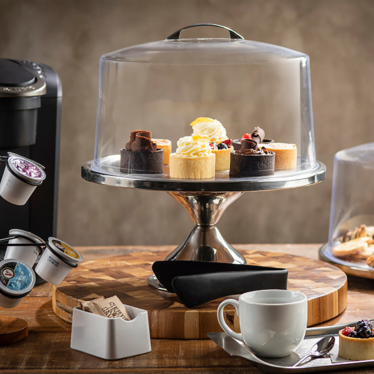 A cake stand with a stainless steel base and clear plastic cover displays assorted mini cakes—chocolate, vanilla, and berry. Nearby are a white coffee cup, coffee pods, sugar packets, and pastries on a wooden surface.