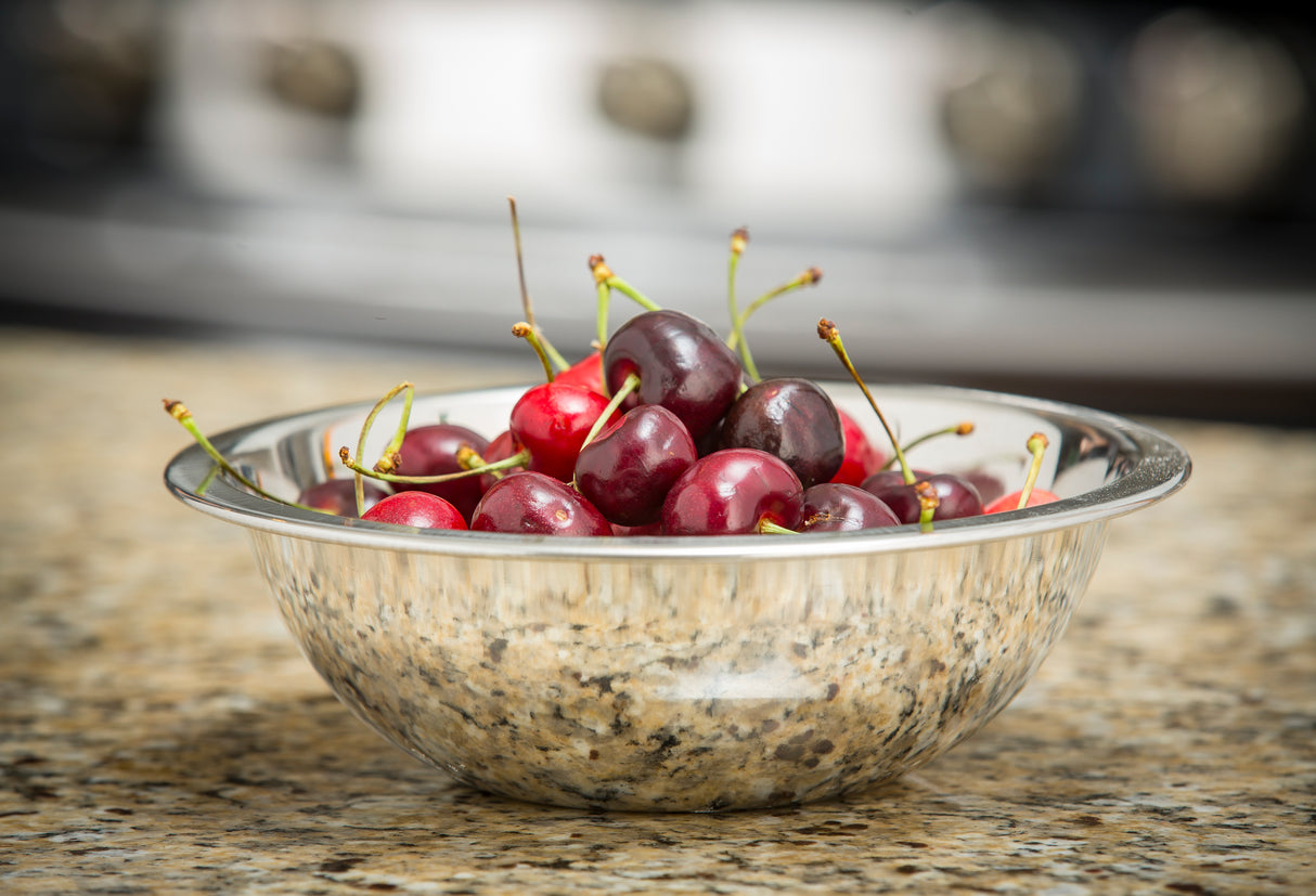 A clear mixing bowl filled with fresh, dark red cherries with green stems sits on a speckled granite countertop. The background is softly blurred, highlighting the cherries and the reflective surface of the 3 qt. stainless steel bowl.