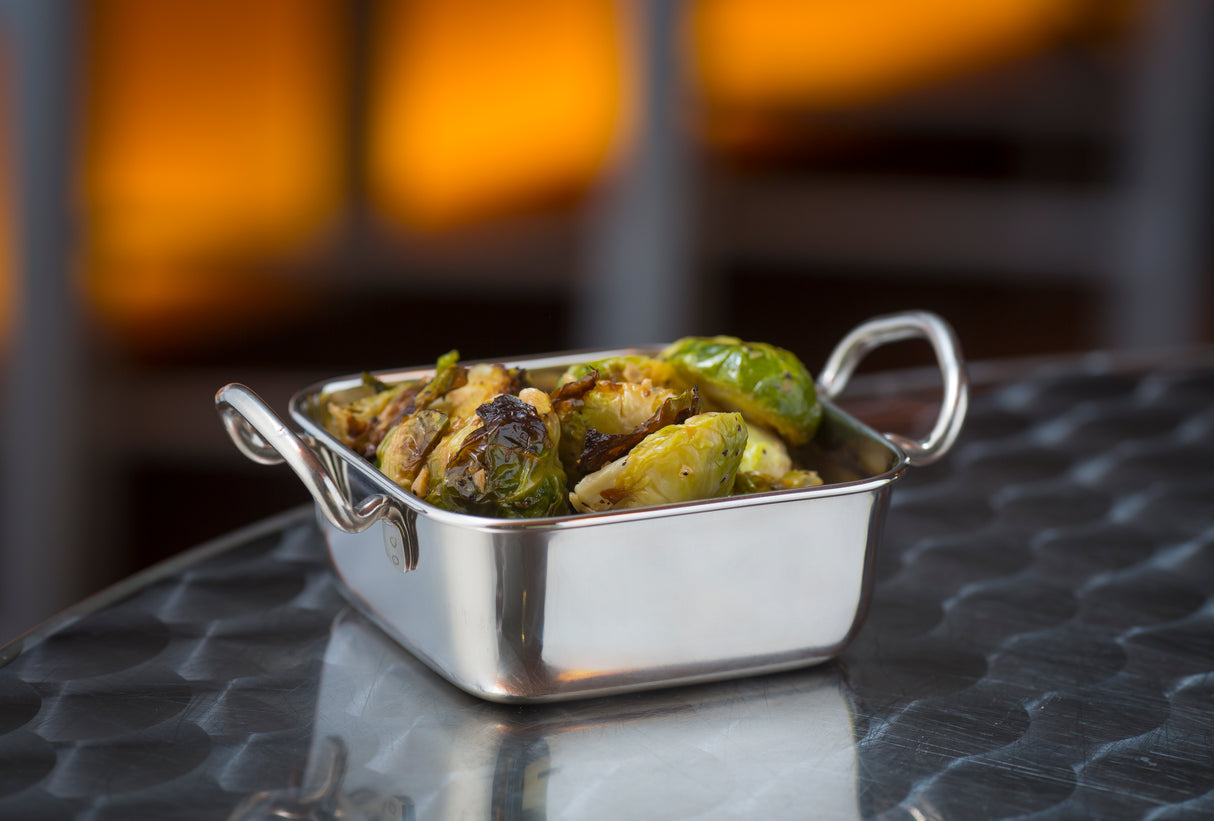 A small stainless steel mini roast pan with handles holds roasted Brussels sprouts, slightly charred and seasoned, on a round metal table. The background is softly blurred with warm orange and brown tones.