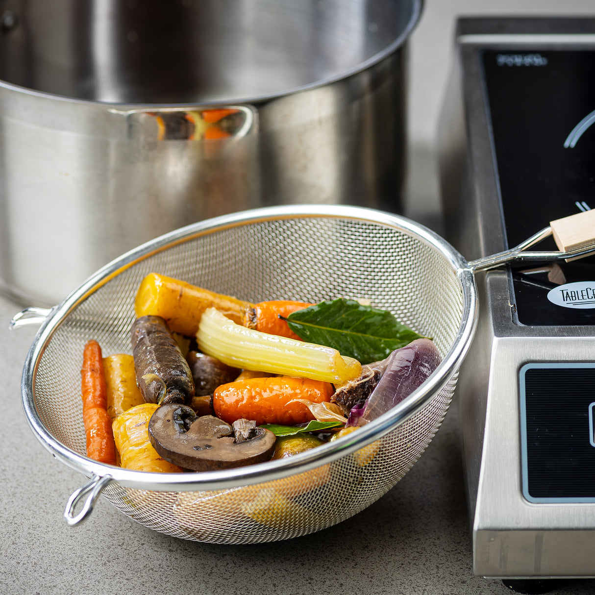 A metal strainer with a wooden handle and medium mesh, filled with colorful cooked vegetables—mushrooms, carrots, celery, and leafy greens—rests on a countertop between a large stainless steel pot and an electric stovetop, suggesting soup-making.
