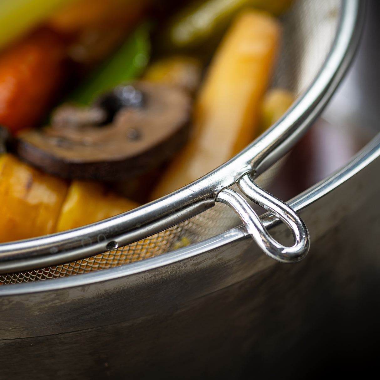 A close-up of a medium mesh strainer with a wooden handle resting on a metal pot, filled with cooked vegetables like mushroom slices, carrots, and green beans. Vibrant colors and shallow depth of field highlight the strainer’s edge and food texture.