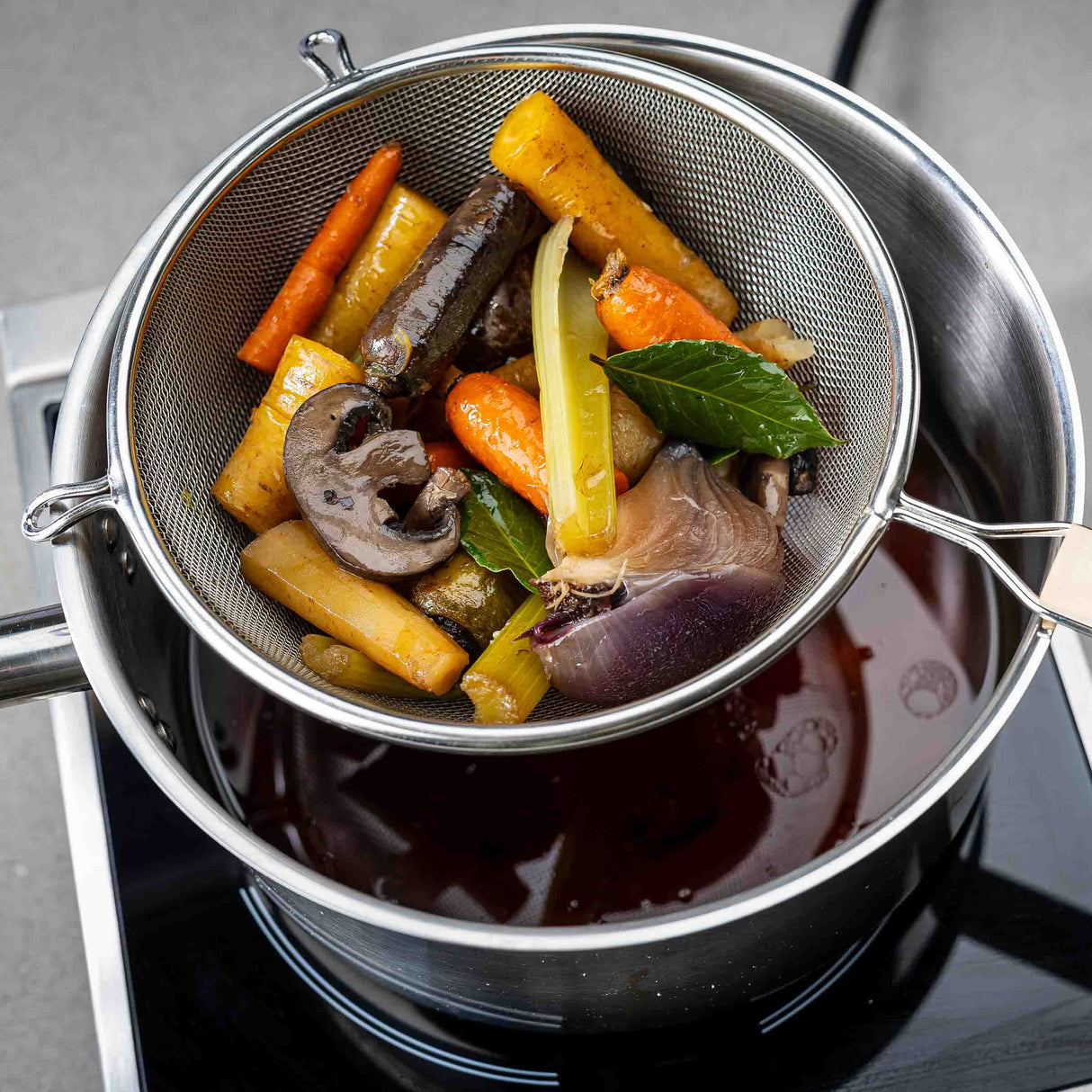 A medium mesh strainer with a wooden handle holds cooked carrots, celery, mushrooms, onion, and bay leaves over a pot of dark broth, draining the vegetables while allowing the flavorful liquid to collect below.