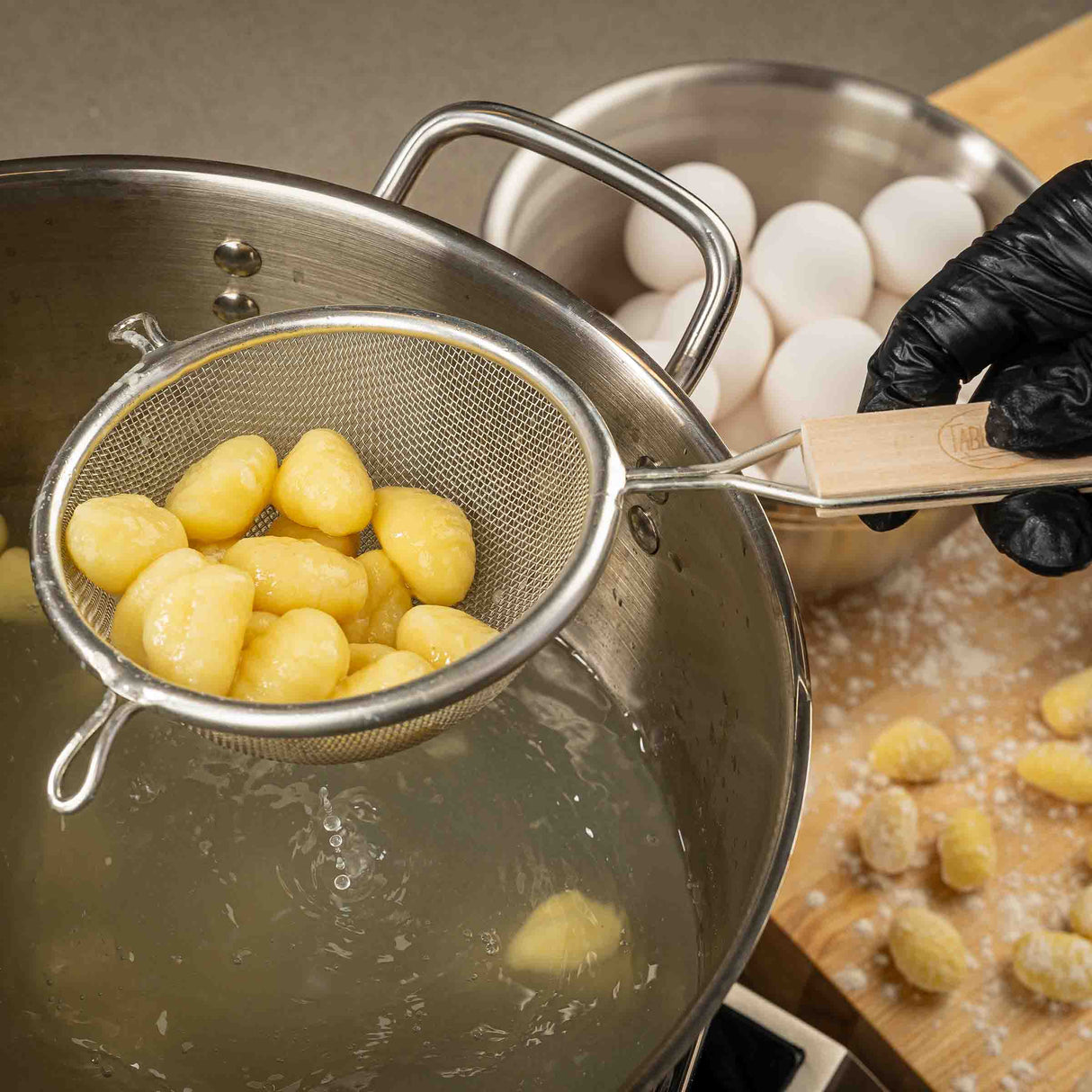 A hand in a black glove holds a fine mesh strainer with cooked gnocchi over a pot of hot water. In the background, eggs and uncooked gnocchi rest on a floured wooden surface as steam rises, highlighting the active cooking process.