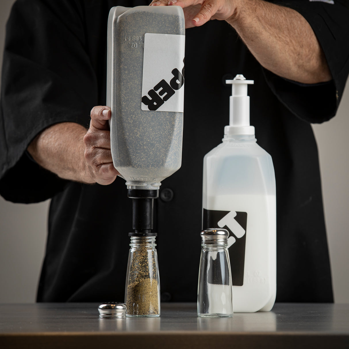 A person in a black shirt uses a pepper refiller and funnel to refill a clear glass shaker. Next to them is a salt container with a pump. Both containers are dishwasher safe, and the shaker’s metal lid rests on the table.
