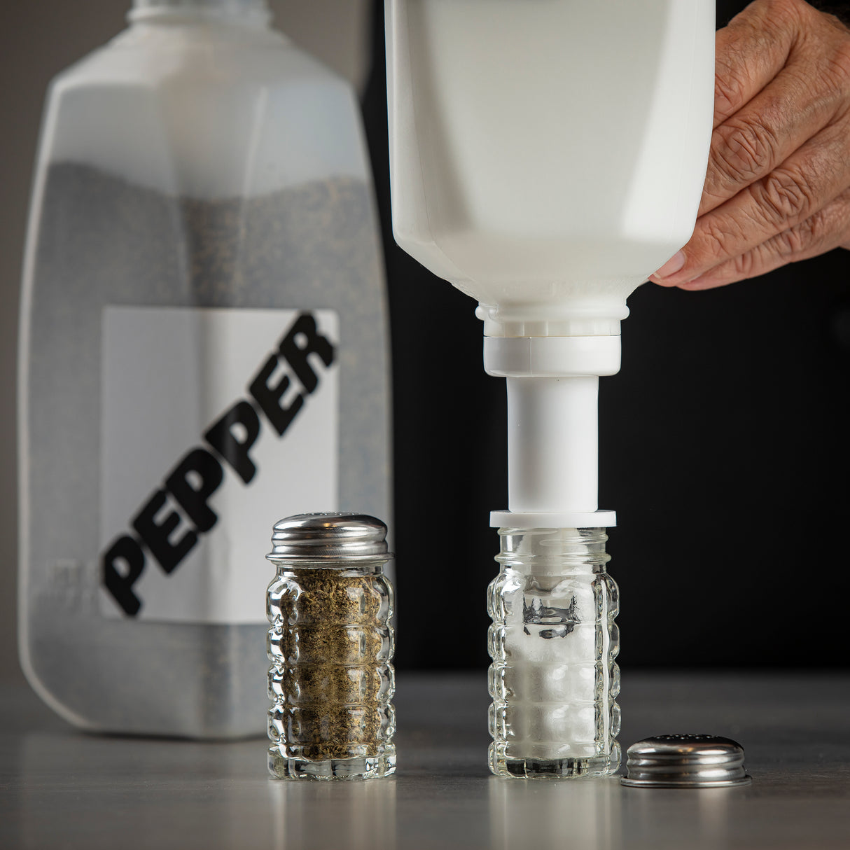 A hand uses a Salt Refiller to pour salt into a glass shaker. A pepper shaker and a large plastic container labeled PEPPER sit nearby. The cap from the salt shaker rests on the gray surface. All items are dishwasher safe for easy cleaning.