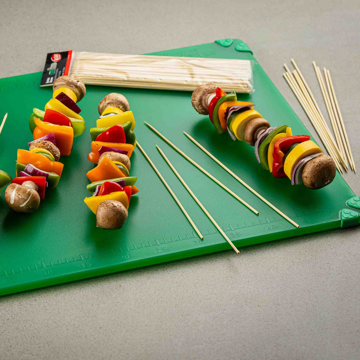 A green cutting board displays TableCraft 10 inch bamboo skewers, some threaded with mushrooms and colorful bell peppers, while others remain empty. Unused bamboo skewers and a packaged bundle rest on the board atop a gray countertop.