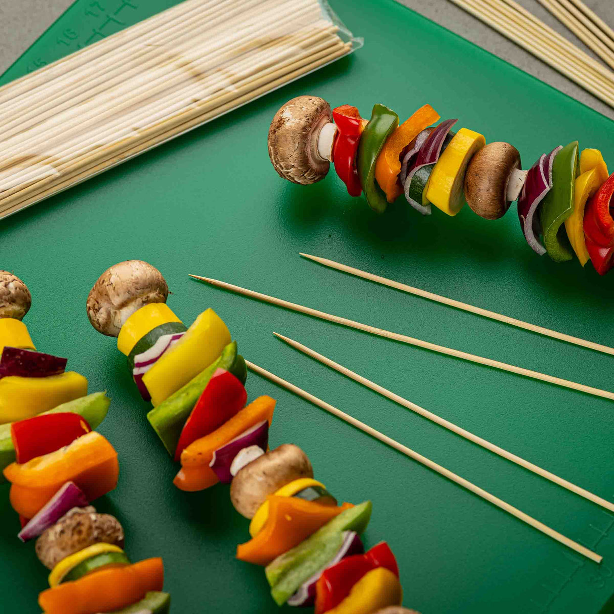 Three 10 inch TableCraft bamboo skewers, threaded with colorful vegetables (bell peppers, red onion, mushrooms), rest on a green cutting board. More empty bamboo skewers and an unopened pack are nearby. The veggies are cut into chunky pieces.