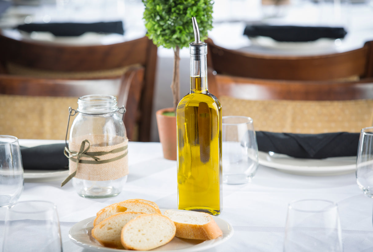 A Prima Bottle of olive oil with a stainless steel pourer sits on a white tablecloth beside sliced bread, a mason jar wrapped with twine, empty glasses, and a small potted plant; chairs are in the blurred background.