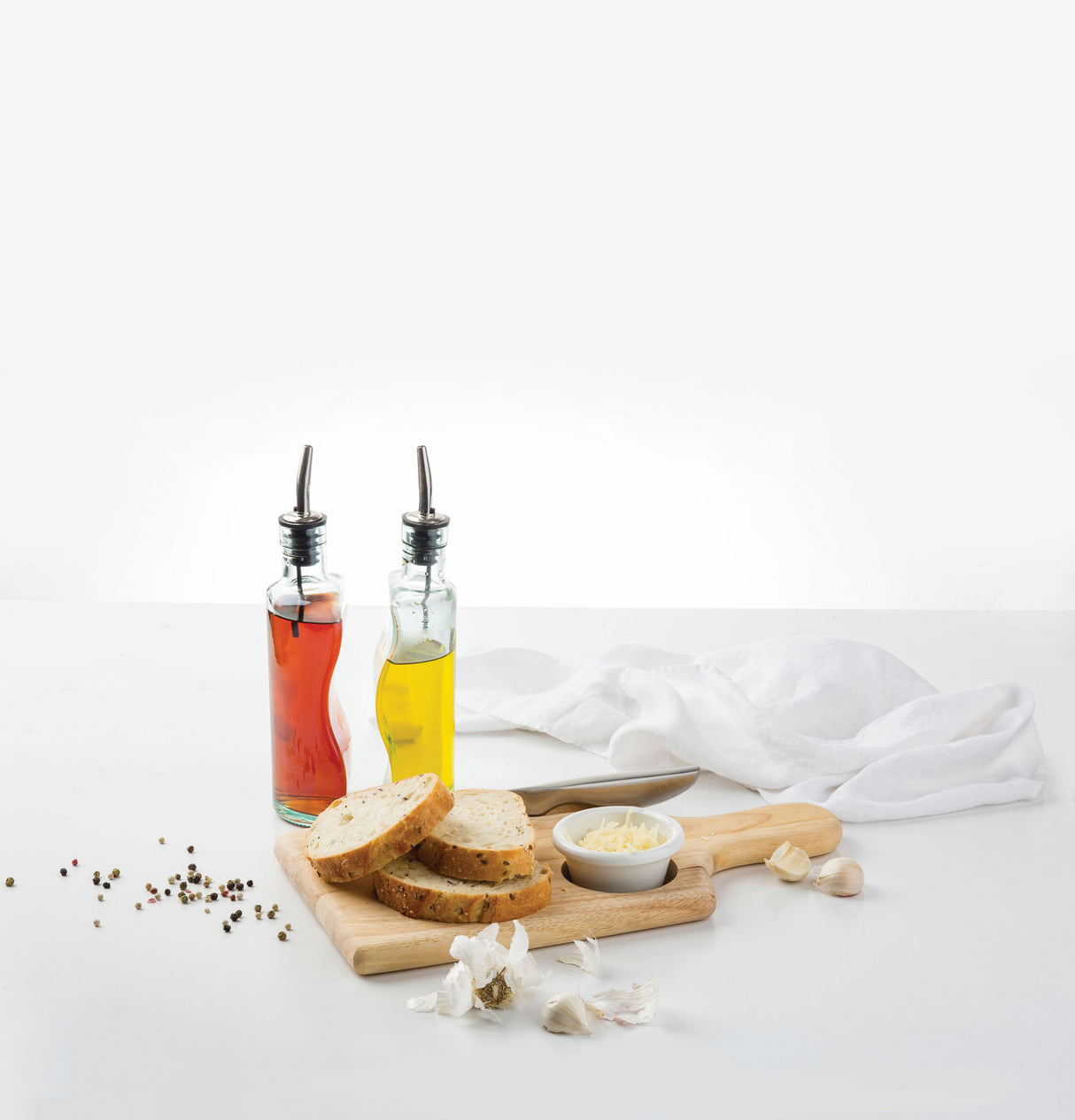 Two green glass bottles of oil and vinegar with stainless steel pourers sit on a white table next to sliced bread, grated cheese, garlic cloves, and scattered peppercorns. The setting is clean and minimal with neutral tones.
