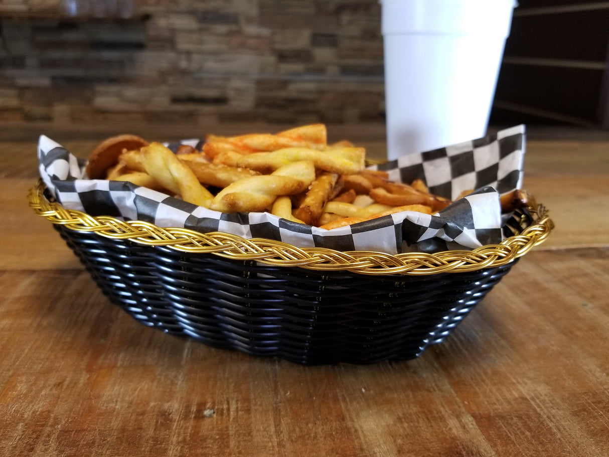 A hand-woven black basket with gold trim, lined with black-and-white checkered paper, holds a serving of seasoned French fries. In the background, a white disposable cup sits on a rustic wooden table against a blurred brick wall.
