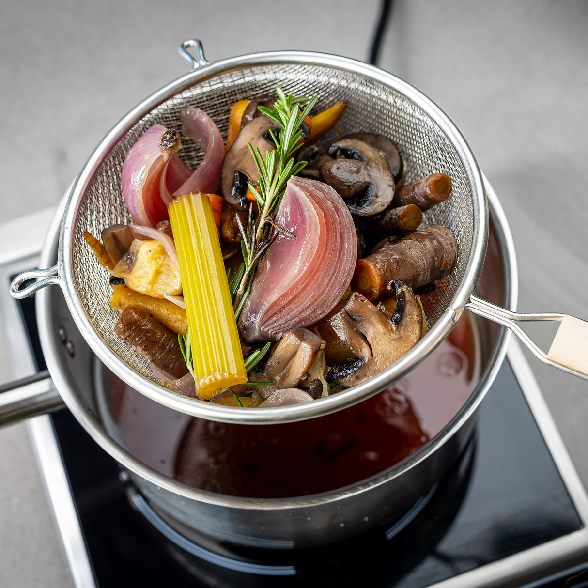 A metal strainer with a wooden handle and double medium mesh holds cooked vegetables—onion, celery, carrot, mushrooms, and rosemary—over a saucepan of brown broth on the stovetop. The simmered vegetables are being freshly drained.