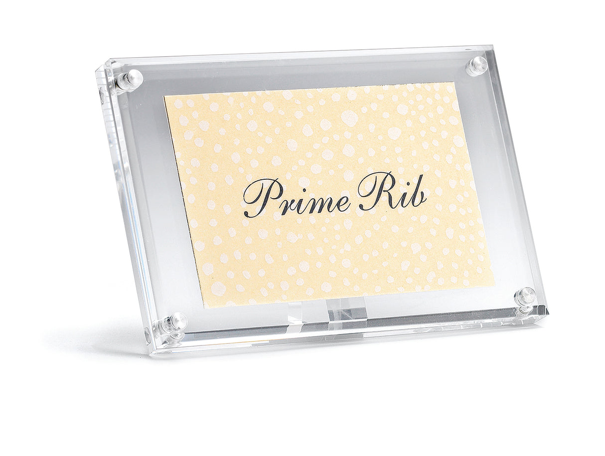 A clear acrylic sign holder displays a cream-colored card with white dots and elegant black script reading Prime Rib. The sign holder stands at a slight angle against a plain white background.