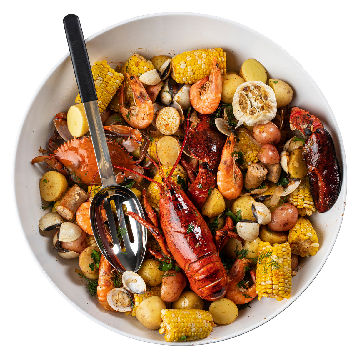 A large white bowl filled with a seafood boil featuring lobster, crab, shrimp, clams, corn on the cob pieces, red potatoes, and halved garlic bulbs. A colorful and rustic dish is served with a stainless steel slotted serving spoon on the side.